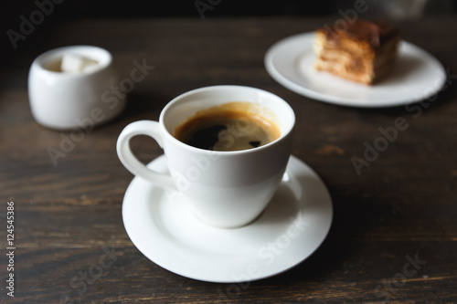 A white cup of black coffee  a piece of cake on the white plate  sugar bowl on the wooden table in a cafe. Selective focus  small depth of fieild.