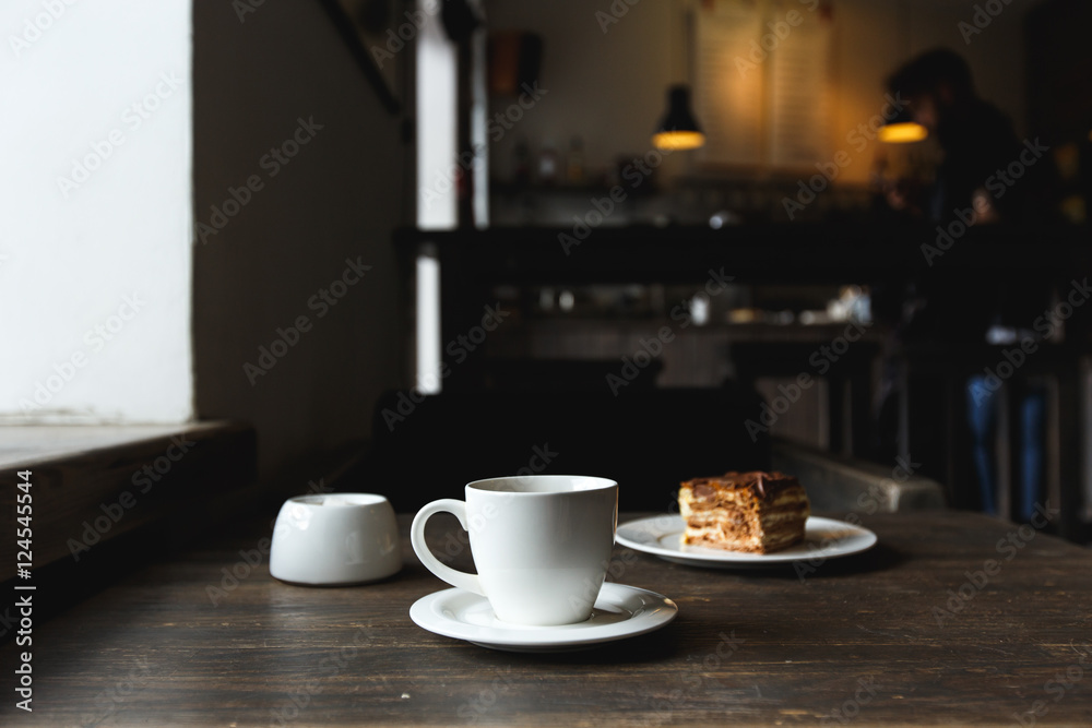 A white cup of black coffee, a piece of cake on the white plate, sugar bowl on the wooden table in a cafe. Selective focus, small depth of fieild.