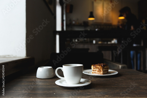 A white cup of black coffee  a piece of cake on the white plate  sugar bowl on the wooden table in a cafe. Selective focus  small depth of fieild.