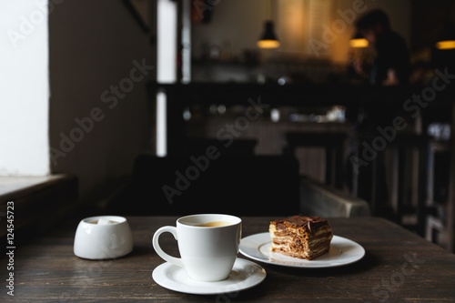 Fototapeta Naklejka Na Ścianę i Meble -  A white cup of black coffee, a piece of cake on the white plate, sugar bowl on the wooden table in a cafe. Selective focus, small depth of fieild.
