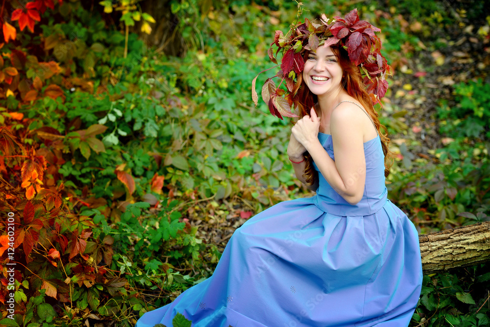 Charming girl in a wreath from autumn leaves, in a blue dress sitting on a log on the background of red and green bushes in a Sunny day and cheerfully laughs. Portrait. Horizontal view