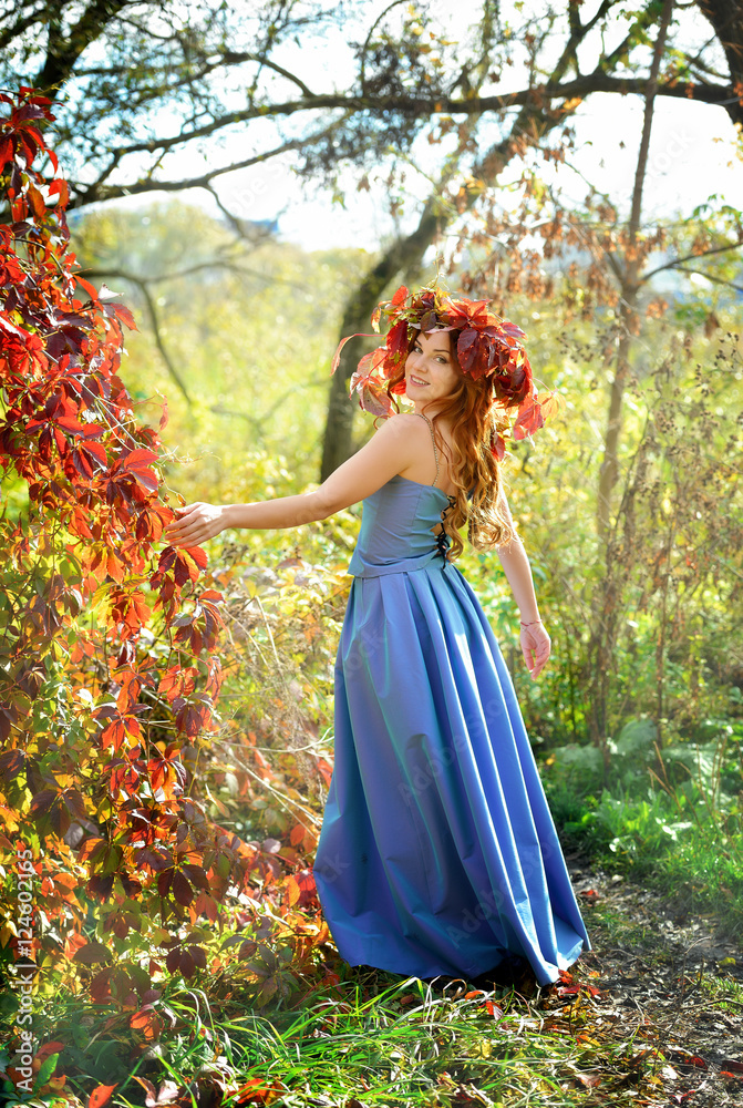 Happy girl in a wreath from autumn leaves, in a blue dress, standing near the red bushes on a yellow background on a Sunny day, posing and smiling. Portrait. Vertical view