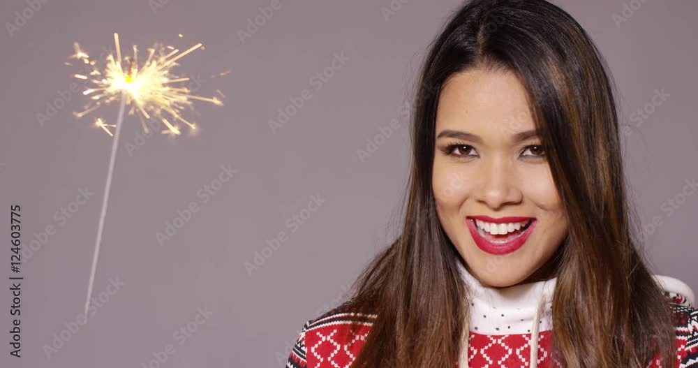 Fun young woman celebrating Christmas with a sparkler watching the fiery sparks with a happy grin in a stylish winter outfit Fun young woman celebrating Christmas with a sparkler watching the fiery sparks with a happy grin in a stylish winter outfit