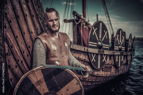 Viking warrior with sword and shield standing near Drakkar on seashore. photo