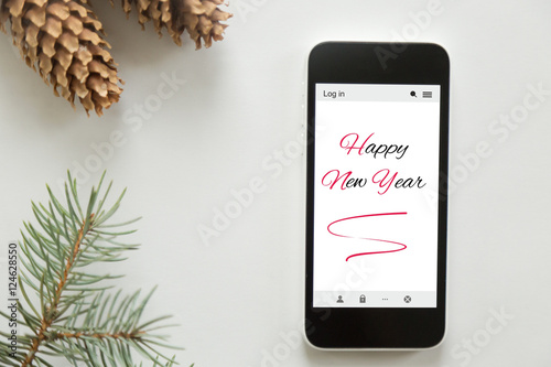 Top view of a mobile phone lying on a white office desk, Happy new year on the screen. Cones, fir tree branch around. New year concept, lay flat