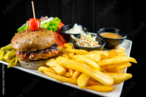 Plate with beef burger, bowl of red, white sauce, golden french fries, pickles and green vegetables. Studio shot with black wooden background.