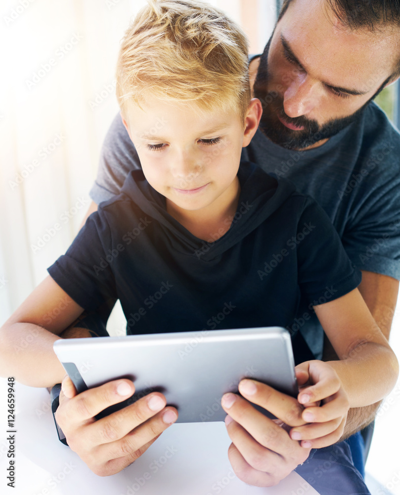 Father and his little son playing together on mobile computer, resting indoor.Bearded man with young boy using tablet PC in sunny house.Vertical, blurred background. Father and his little son playing together on mobile computer, resting indoor.Bearded man with young boy using tablet PC in sunny house.Vertical, blurred background.