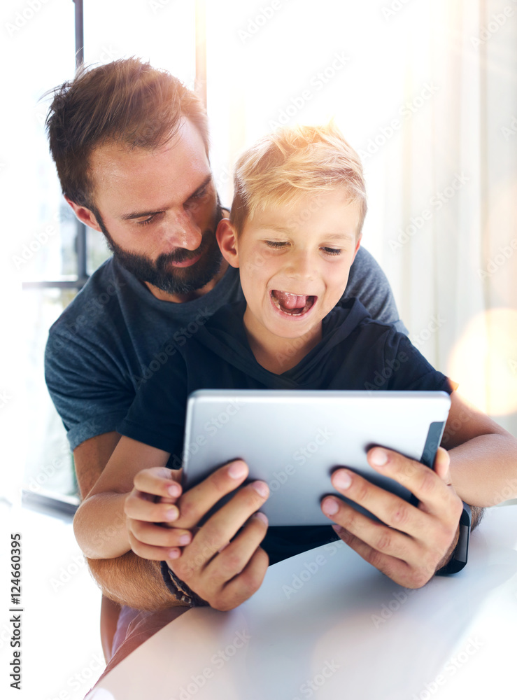 Young boy sitting with father at the table and using pc tablet in modern loft. Vertical, blurred background. Sunlights effect. Young boy sitting with father at the table and using pc tablet in modern loft. Vertical, blurred background. Sunlights effect.