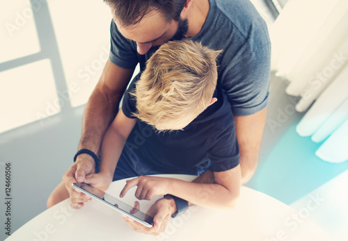 Young boy sitting with father and touching screen of pc tablet in sunny modern loft. Horizontal, blurred background. Top view,sunlights effect.