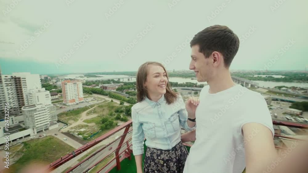A guy brought his girlfriend on the roof to make a surprise. She opened her eyes and smiles. Man takes them to the camera. Selfi. An unexpected gift. Nice view of the city from the roof. A guy brought his girlfriend on the roof to make a surprise. She opened her eyes and smiles. Man takes them to the camera. Selfi. An unexpected gift. Nice view of the city from the roof.
