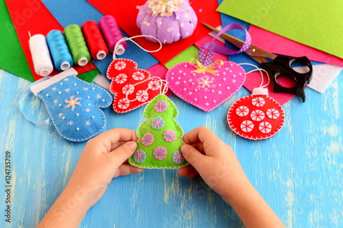 Small child holds felt Christmas tree in his hands. Child shows Christmas crafts. Felt fur-tree, mitten, heart, ball decor on wood background. Flat felt sheets, scissors, sewing set, pins, pincushion
