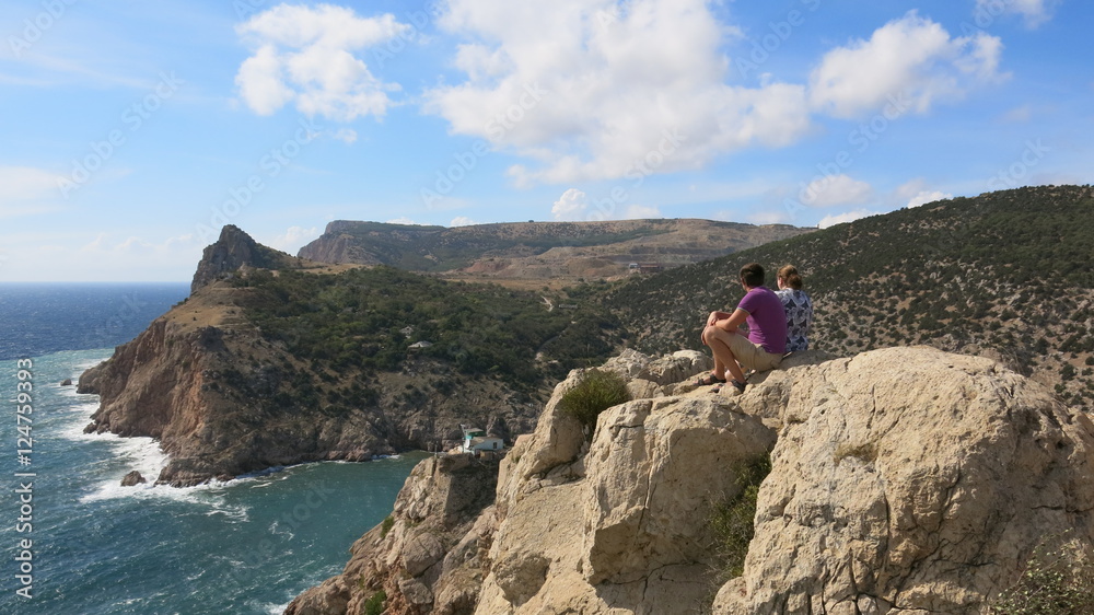 Two people sitting on the edge of a cliff and look down on the sea