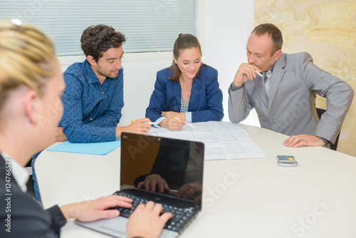 Three people in meeting, secretary using laptop