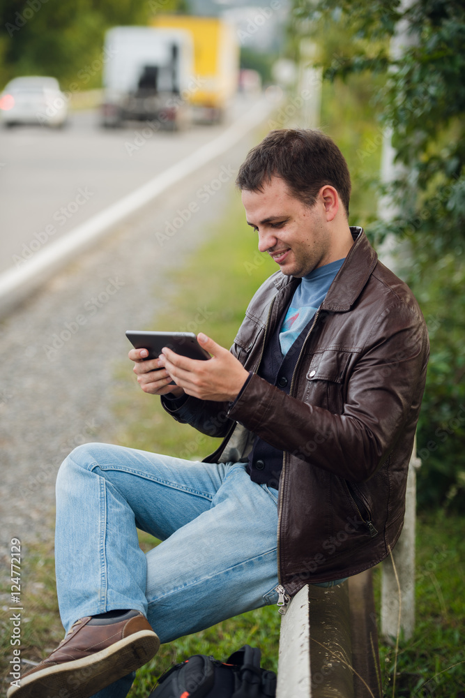 Man orders taxi from his tablet sitting on a road fence Man orders taxi from his tablet sitting on a road fence