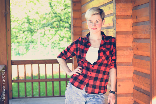 Fashionable portrait of a young beautiful woman in red lumberjack shirt, jeans, white t-shirt and sneakers. Trendy blonde hipster girl having fun on a terrace of a rich wooden house on a sunny day.