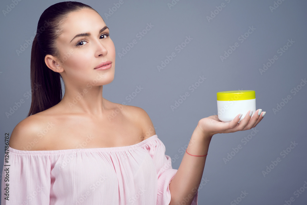 Portrait of young beautiful brunette with ponytail holding cosmetic jar on her palm and smiling. Cosmetics and body care concept. Copy-space. Studio shot