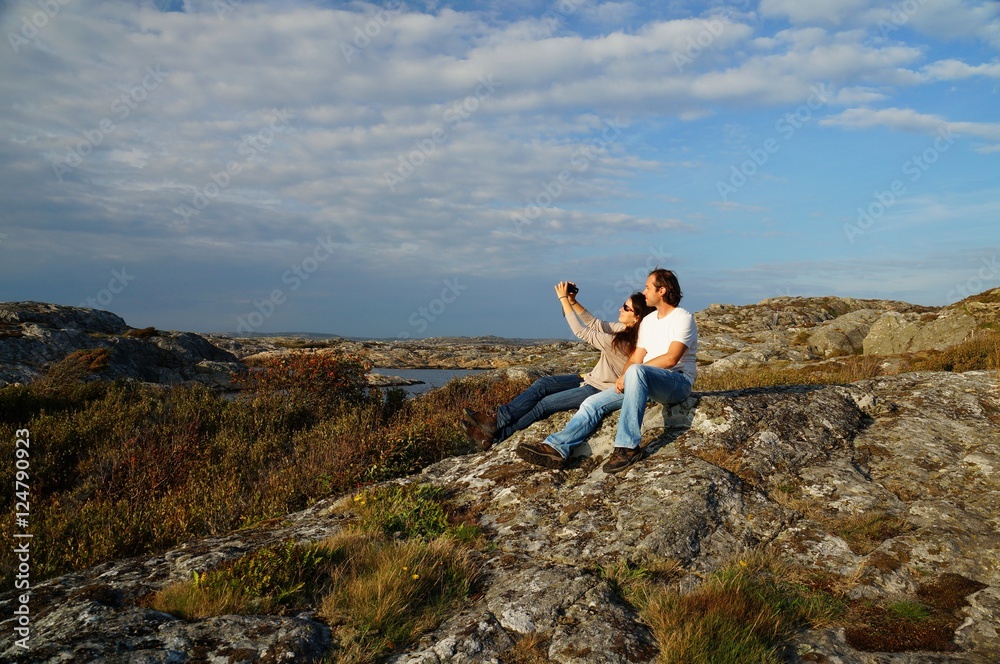 Obraz premium Couple taking a selfie on large stone covered with colorful lichens at a coastal heathland at a hiking path next to Marstrand, Kungälv Municipality, Västra Götaland County, Sweden