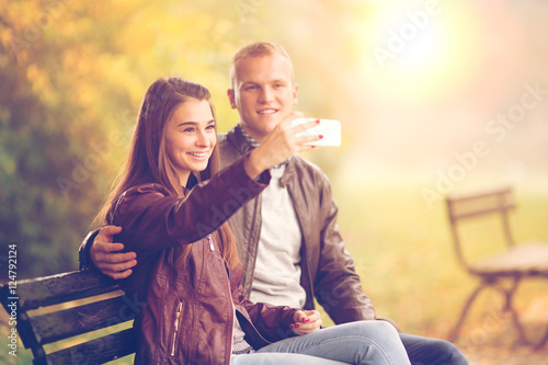 Young couple are taking photo of themselfs with they mobile phone on a beautiful Autumn day. They are sitting on a bench and smiles. photo
