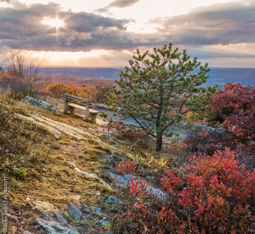 A lone bench overlooks beautiful fall foliage under a majestic setting sunburst at the top of New Jersey at High Point State Park
