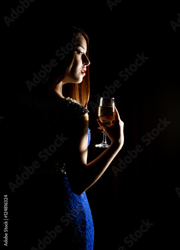 Young celebrating woman in a blue dress holding a glass of champagne on a dark background. play of light and shedow photo