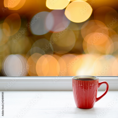 Cup of coffee on the window sill. In the background the bright lights of the city as a Christmas background
