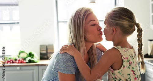 Mother and daughter kissing and hugging at home in bright kitchen. Sunlight coming through window