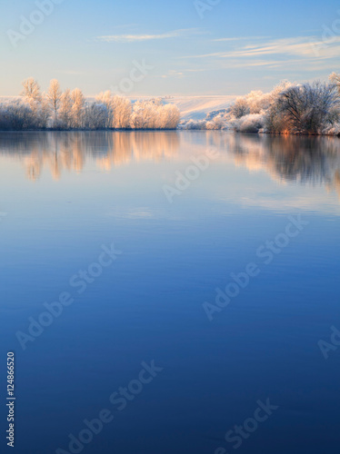 Winter Landscape Reflecting in River at Sunrise, Trees covered by hoarfrost and snow, blue sky, space for text