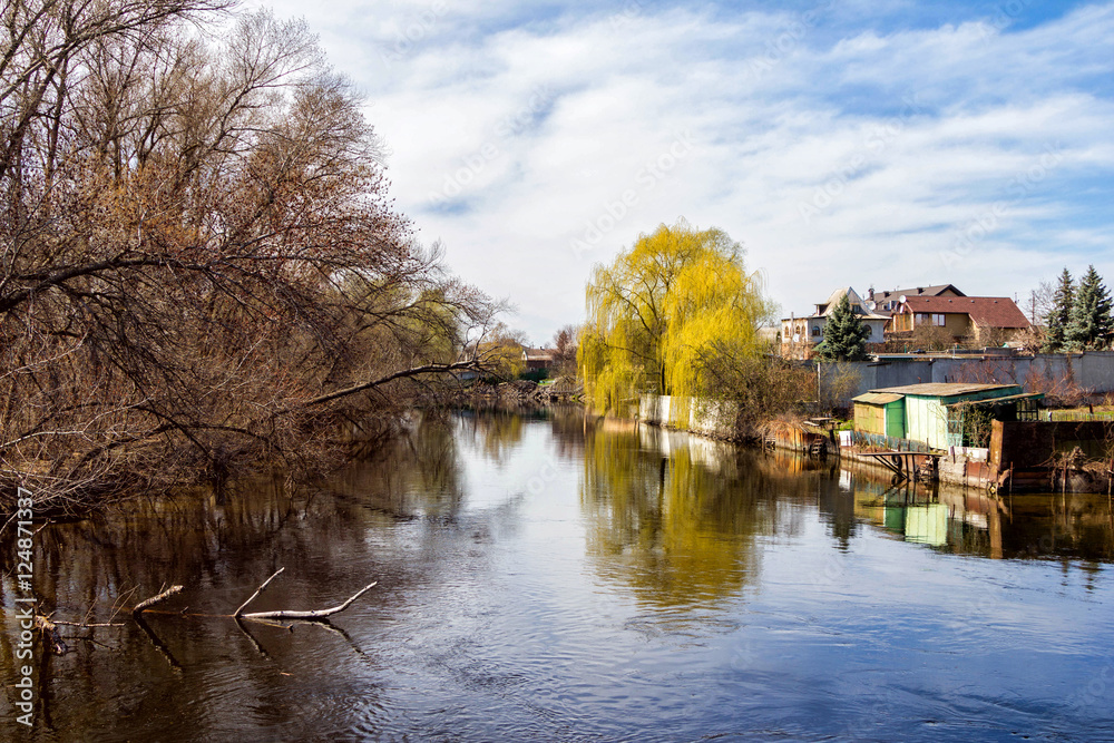 Springtime landscape - river with small village and forest on the banks, dry tree branches floating in a water with sky reflections Springtime landscape - river with small village and forest on the banks, dry tree branches floating in a water with sky reflections