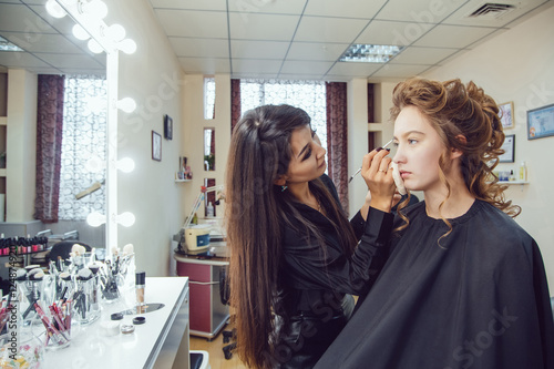 Make up artist doing professional makeup of young woman near the mirror in beauty studio