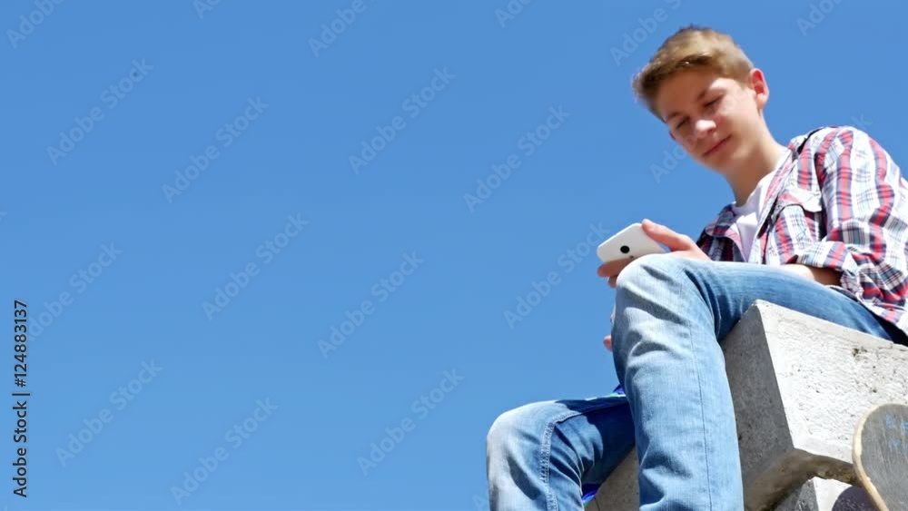 Low angle view of teenager boy writing sms, texting on smartphone against blue sky Low angle view of teenager boy writing sms, texting on smartphone against blue sky