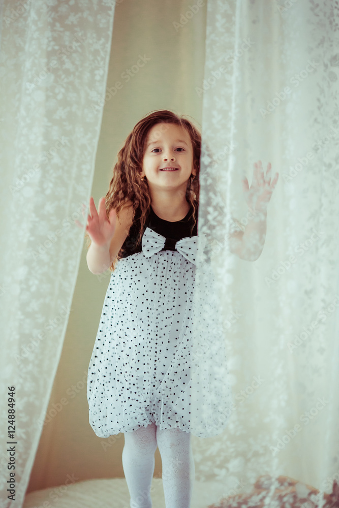 Little curly girl in spotted dress stands behind the white curta