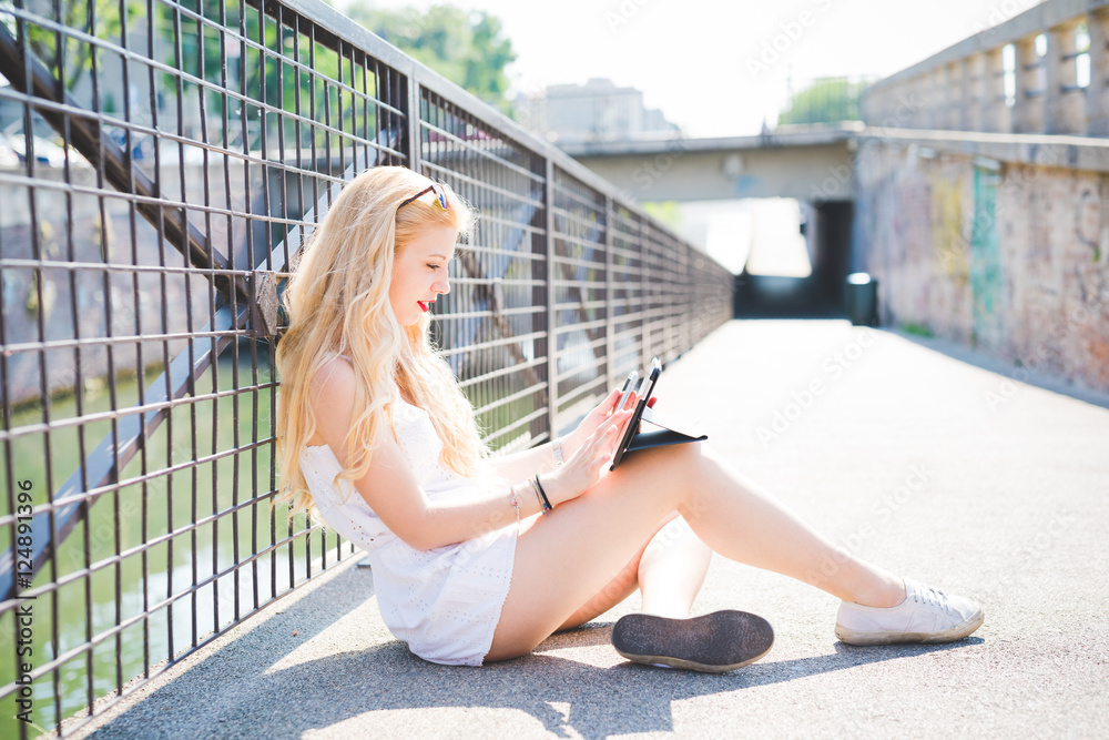 Fototapeta premium young handsome caucasian long blonde hair woman sitting on the floor talking smart phone and using tablet, looking downward the screen, tapping - technology, social network, communication concept