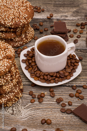 Small white cup of coffee, roasted coffee beans, cookies with sesame seeds, slices of chocolate on wooden background