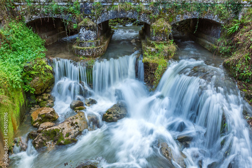 Small waterfall at mountain river under the old bridge in Asturias, Spain. Waterfalls are commonly formed in the upper course of the river. 