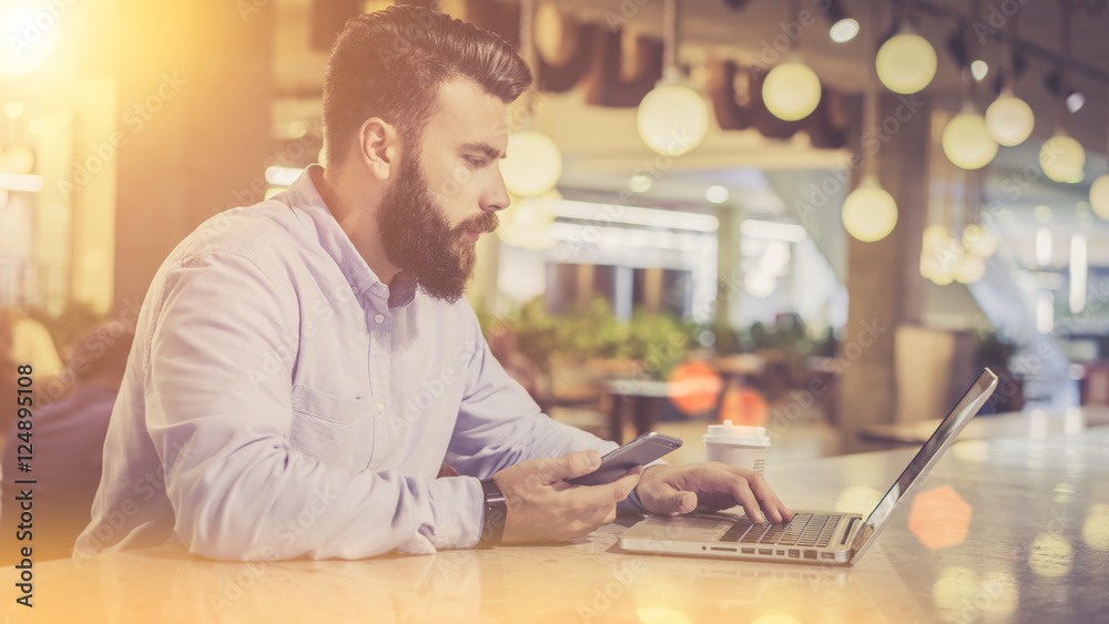 View from the side, a bearded man in a shirt using a laptop sitting at a table in a café, in a man's hand cell phone, on the table cup of coffee.Film effect, blurred background.A man checks the email. View from the side, a bearded man in a shirt using a laptop sitting at a table in a café, in a man's hand cell phone, on the table cup of coffee.Film effect, blurred background.A man checks the email.