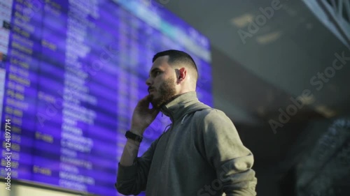 The young man is tapping number on mobile phone standing next to the information board at the airport.