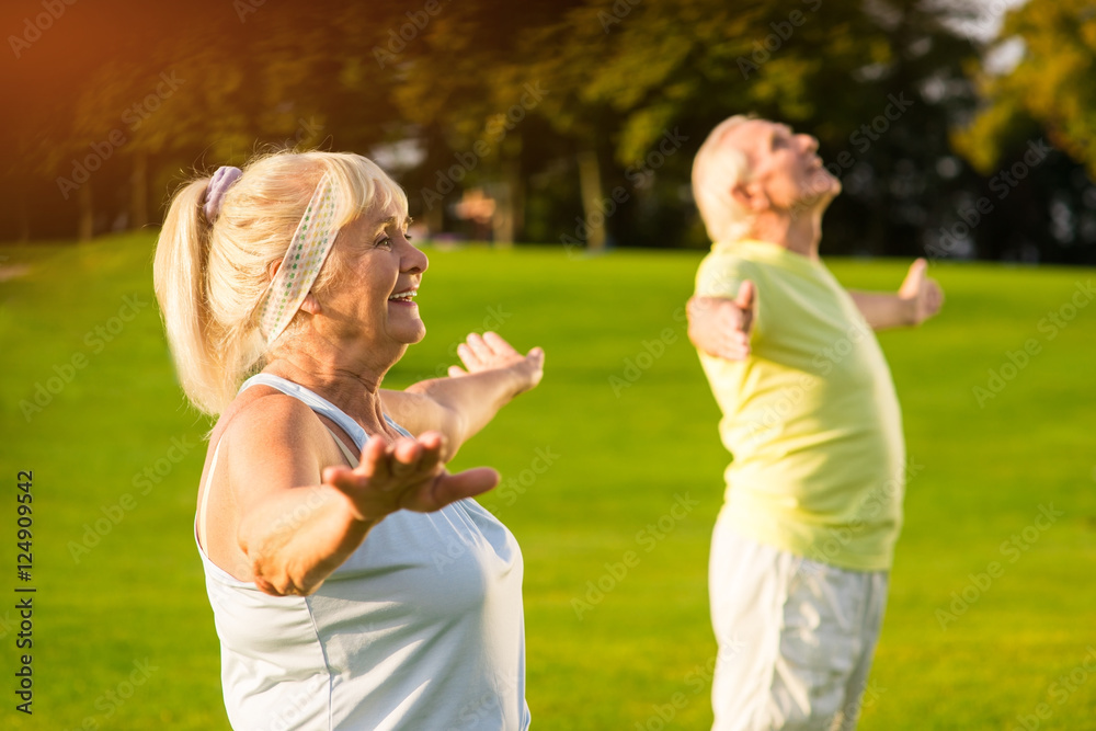 Fototapeta premium Senior woman with outstretched arms. Smiling lady and man outdoors. Gymnastics improves health. Happy and energetic.