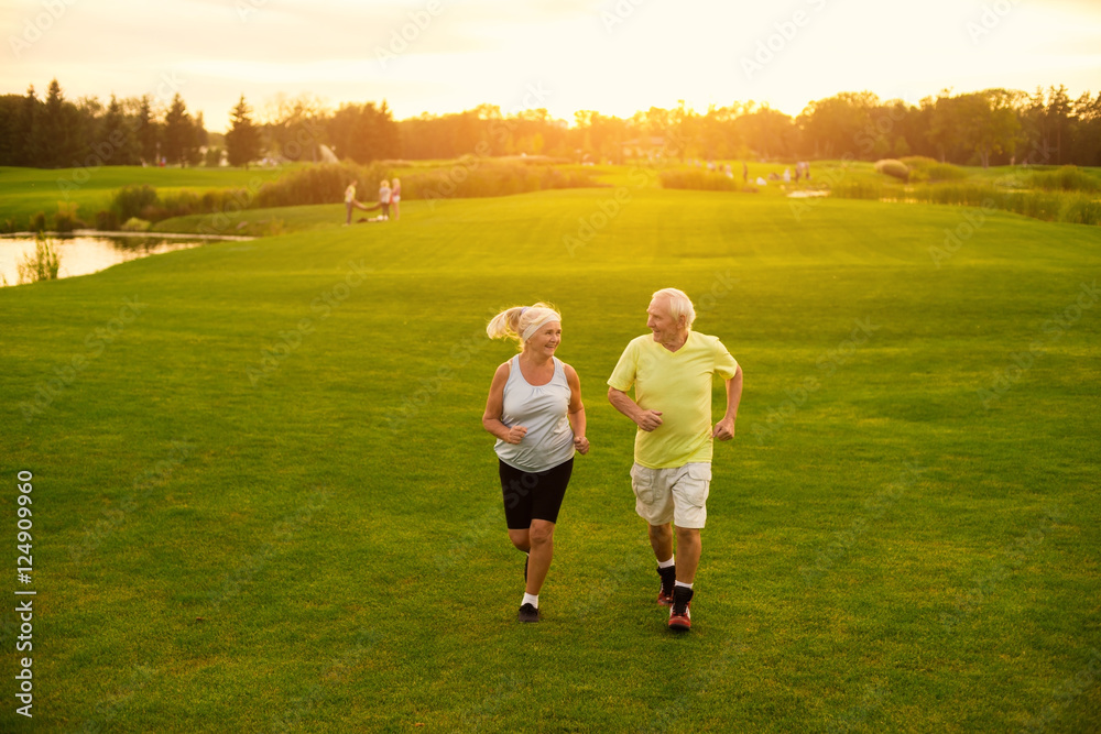 Elderly couple is jogging. Smiling people on meadow. Be in motion. The lightness of moves.