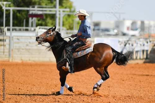 The side view of a rider in cowboy chaps, boots and hat on a horseback performs an exercise during a competition.