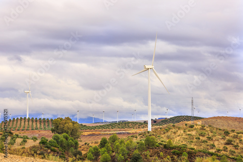 Wind farm in mountainous landscape with cloudy sky, with pines and other trees, also olive groves and other farmlands at background