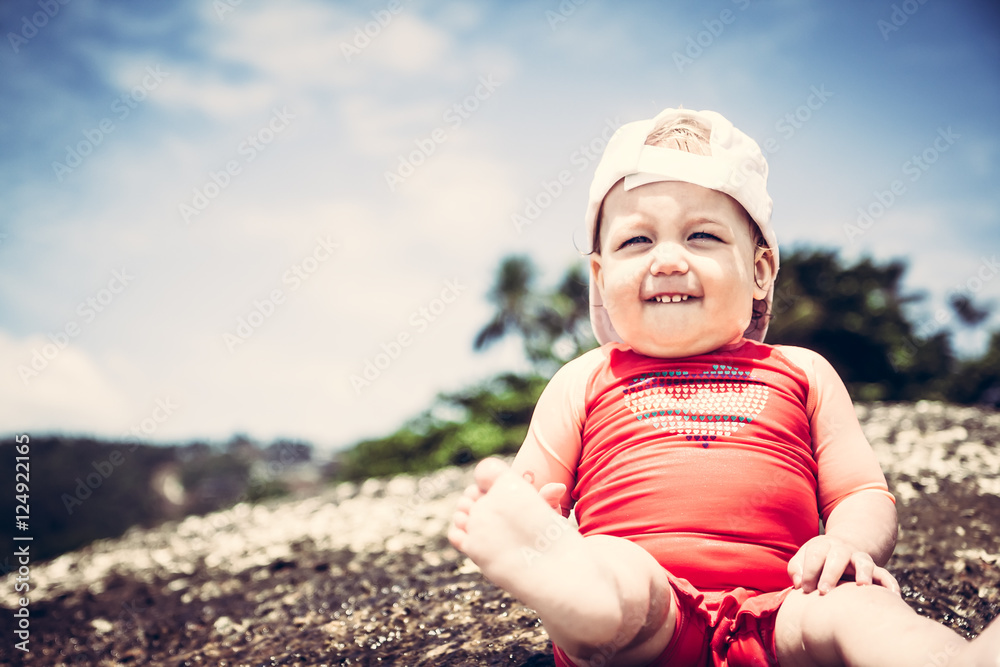 Cheerful baby in sun protective suit on beach looking forward with bright blue sky background and copy space Cheerful baby in sun protective suit on beach looking forward with bright blue sky background and copy space