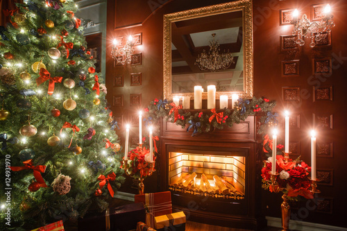 Christmas evening by candlelight. classic apartments with a white fireplace, decorated tree, sofa, large windows and chandelier.