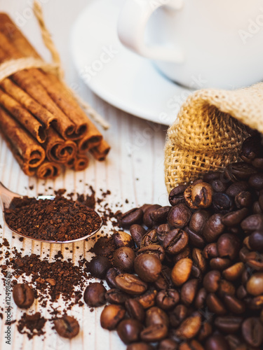 Cup of coffee  pouch with coffee beans  cinnamon sticks and spoonful of ground coffee on the wooden table. Shallow focus