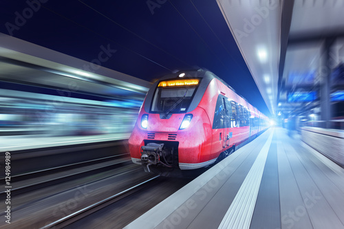 Modern high speed red passenger train moving through railway station at night. Railway station in Nuremberg. Railroad with motion blur effect. Industrial concept . Railroad travel, railway tourism