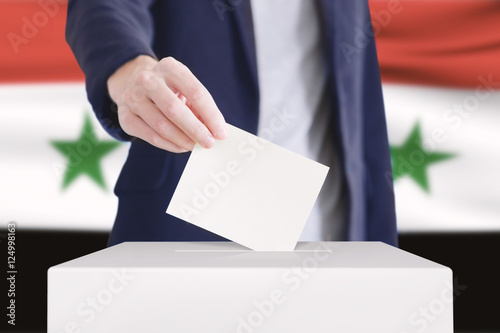 Voting. Man putting a ballot into a voting box with Syrian flag on background.