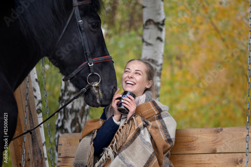 girl in a plaid with a black horse in the autumn under a birch tree on a bench