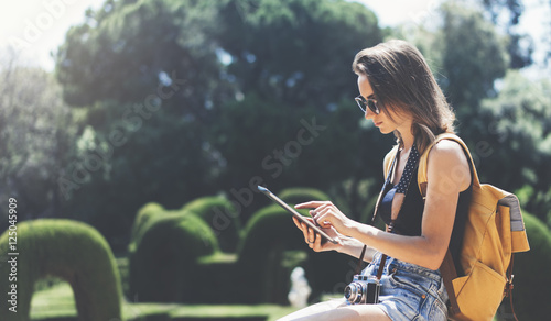 Hipster young girl with backpack and vintage camera using tablet or holding gadget, travel plan. View tourist traveler with sunglasses on background Parc Laberint Barcelona. Mockup for text message photo