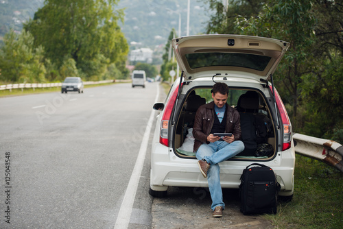 leisure, road trip, travel and people concept - happy man searching location using tablet with online map sitting on trunk of hatchback car outdoors