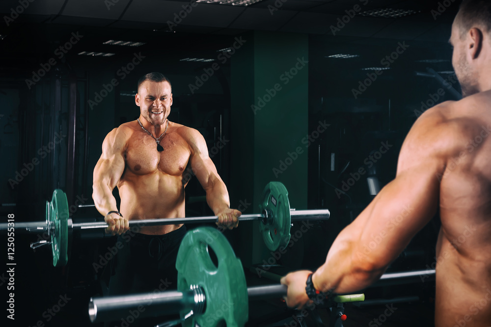 power athletic guy bodybuilder working out biceps with barbell in front of mirrors, in dark gym power athletic guy bodybuilder working out biceps with barbell in front of mirrors, in dark gym