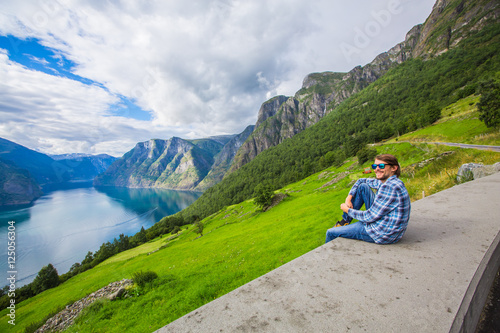 Picturesque Norway mountain landscape. Young man enjoying the view near Geiranger fjord, Norway. Sitting on a cliff. Tourist in blue shirt and sunglass. Copy space. Place for advertising inscription. © nataliakabliuk